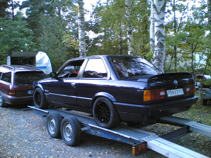 BMW E30 på en släpvagn med träd och annan bil i bakgrunden.