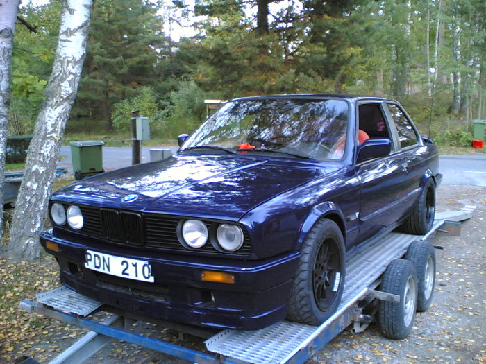 BMW E30 i blått parkerad på en transportsläp i en skogsmiljö.