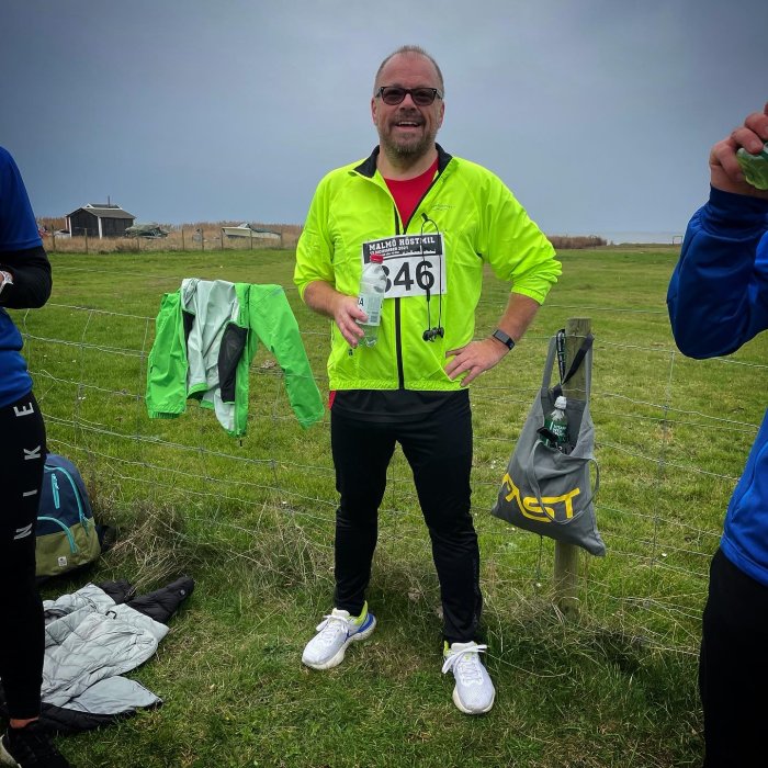 Leende man i löparkläder med nummerlapp står på en gräsmark med sportutrustning i bakgrunden.