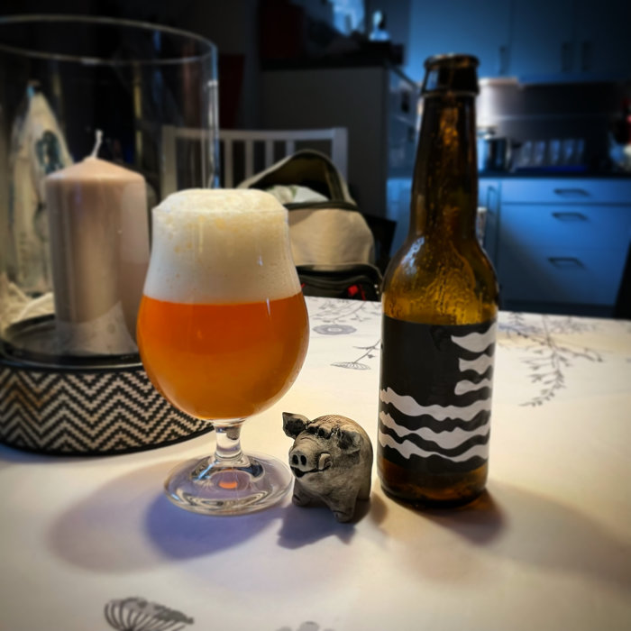Ölglas med skum topp bredvid öppnad flaska och liten grisfigur på bord.