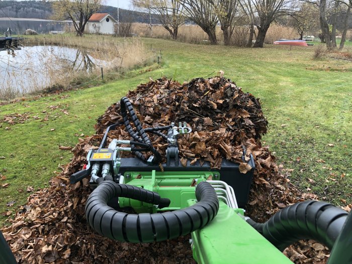 Grönt fordon framför en stor komposthög med löv i en trädgård vid vattnet.