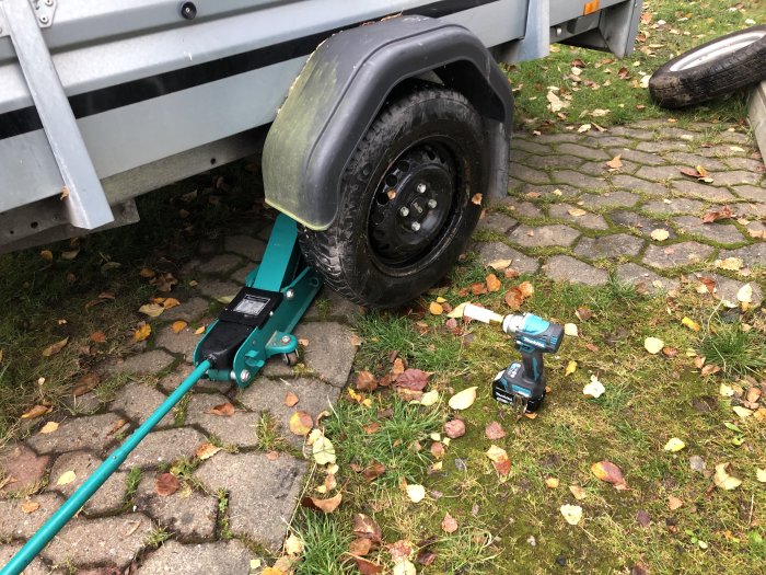 Släpvagn upplyft på domkraft vid hjulbyte med mutterdragare på marken och löv på gräset.