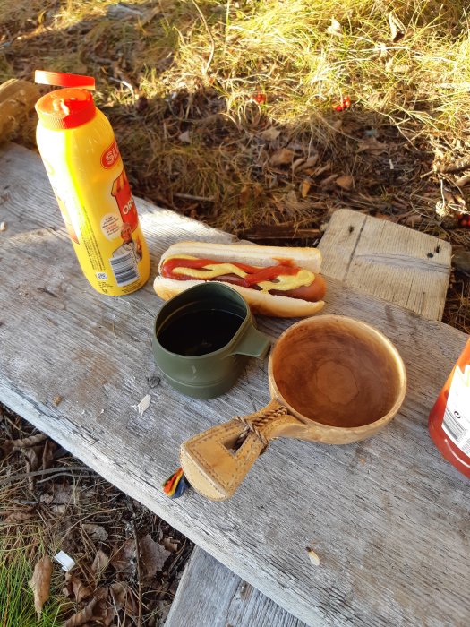 Utomhusfika på en träbänk med korv med bröd, ketchupflaska, kaffemugg och träslev.