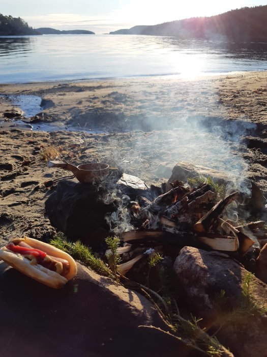 Lägereld vid strandkanten med rök som stiger och en korv med bröd i förgrunden, sjö och skog i bakgrunden.