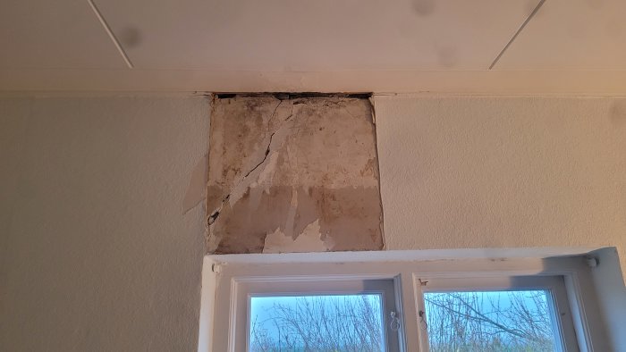 Skadad innervägg med synliga sprickor och mögelskador ovanför ett fönster i ett äldre hus.