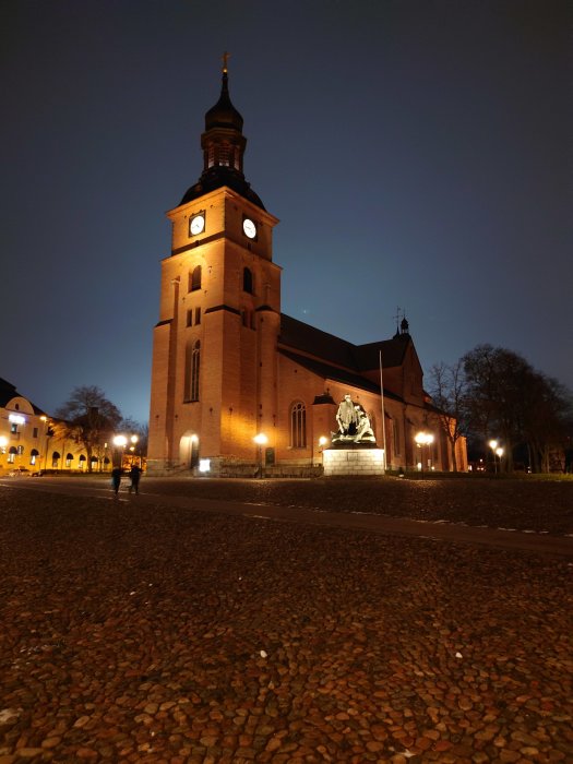 Kyrka belyst på kvällen med staty framför och två personer som promenerar på gatsten.