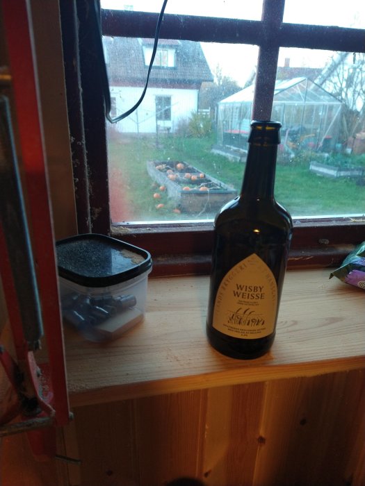 Flaska öl på fönsterbräda framför fönster med utsikt över trädgård med pumpor och växthus, rondeller i burk i förgrunden.