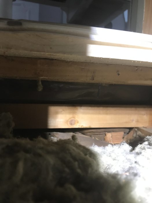 Vy underifrån av ett golv på en övervåning med synliga träbjälkar och gammal mineralull för isolering.