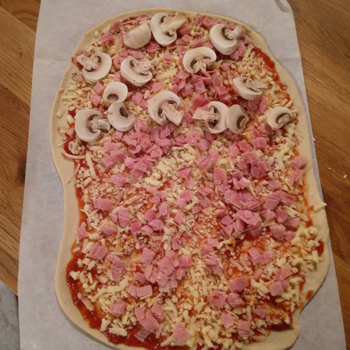 Ogräddad pizza med tomatsås, ost, skinka och champinjoner på bakplåtspapper.