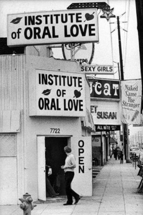 Skyltar med texten "INSTITUTE of ORAL LOVE" och "OPEN" över byggnadens ingång där en person står.