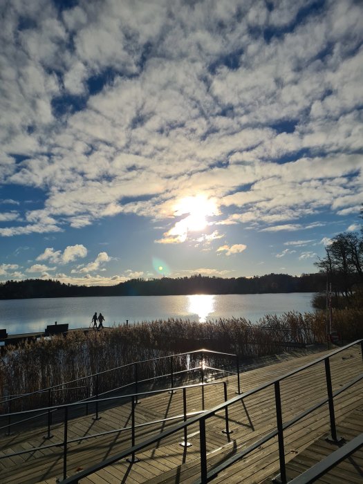 Två personer promenerar på en brygga vid en sjö en solig dag med disiga moln på himlen.