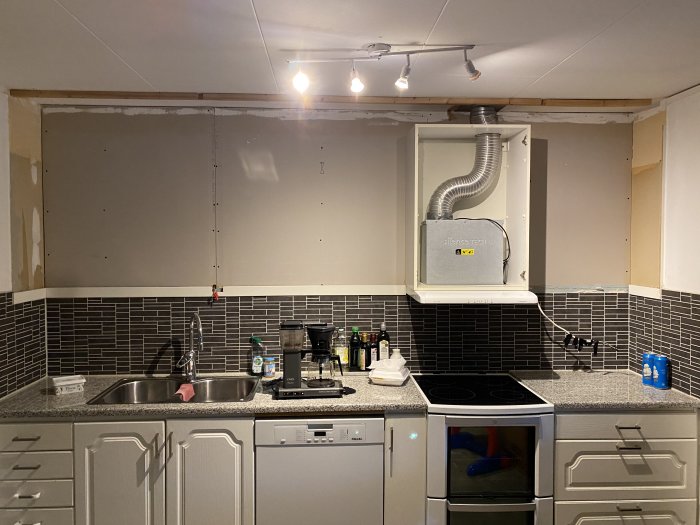 Kök under renovering med omonterade överskåp och synlig ventilation, svart kakel och grå skåp.