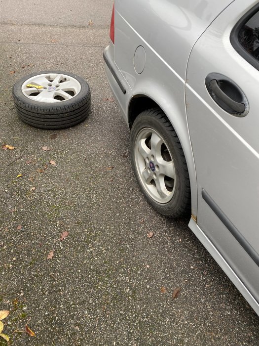 Bild på bil med ett däck som är avmonterat och placerat bredvid bilen på asfalterad mark.