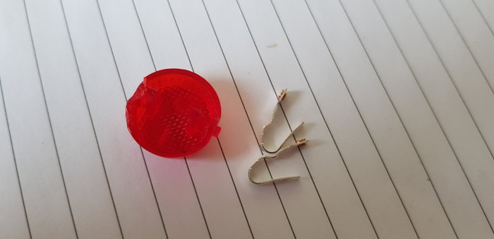 Sönderbruten röd plastknapp och lösa metallstift på vit linjerad bakgrund, komponenter till handdukstork.