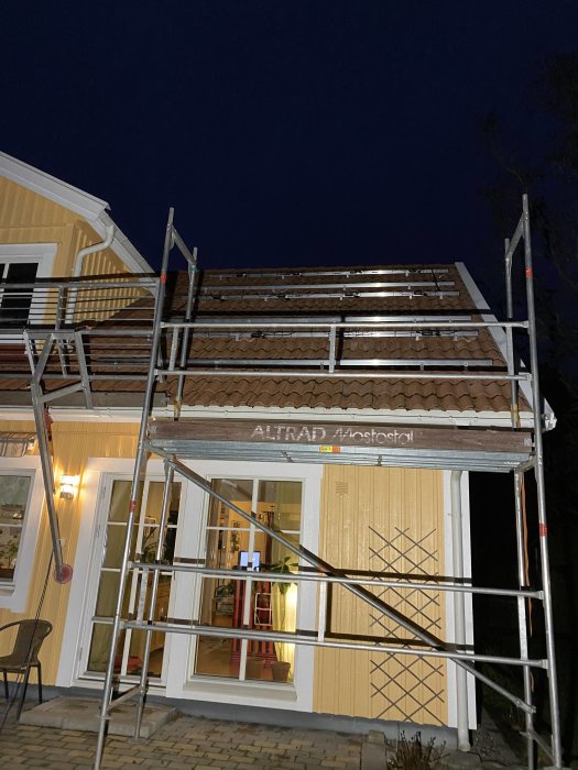 Byggställning framför gult hus med takarbete under kvällshimmel.