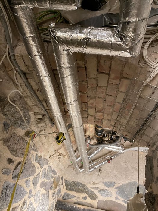 Ventilationsrör med isolering i ett källarrum under renovering, med tegelväggar och stengolv.