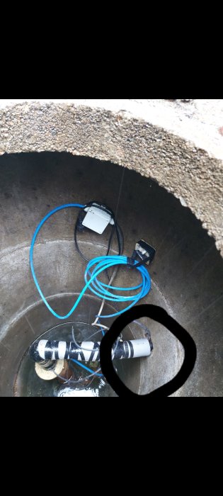 Vy inne i en brunn med rör och kablar, vattenläckage markerat med en svart ring på bilden.