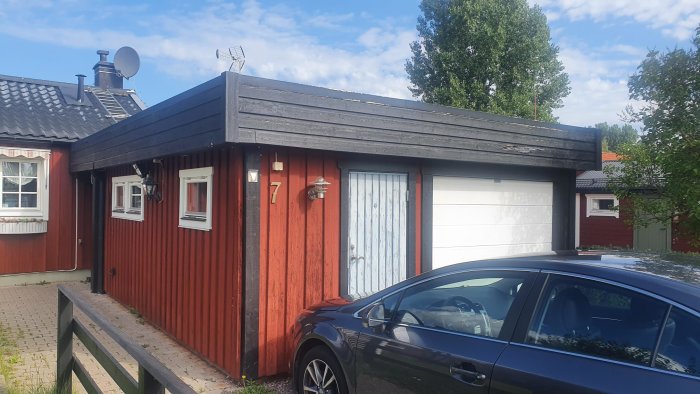 Garage med slitna svarta takplattor bredvid hus med rödfärgad träpanel och vit bil framför.