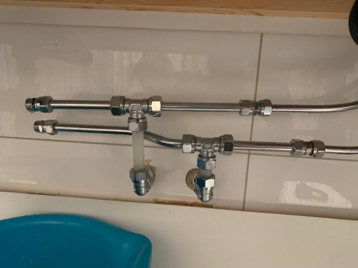 Röranslutningar under handfat med olika kopplingar och en blå hink under.