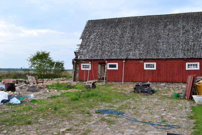 Rödmålad lada med mossa på taket, med byggmaterial och verktyg utspridda på gården.