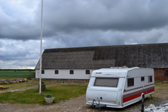 En campingvogn står foran en hvid landejendom med mosbegroet tag under en skyet himmel. En stige læner op ad bygningen.