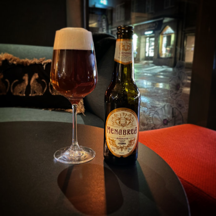 Öppnad ölflaska och ett glas fyllt med öl på ett bord inne på en bar, med en suddig stadsgata i bakgrunden.