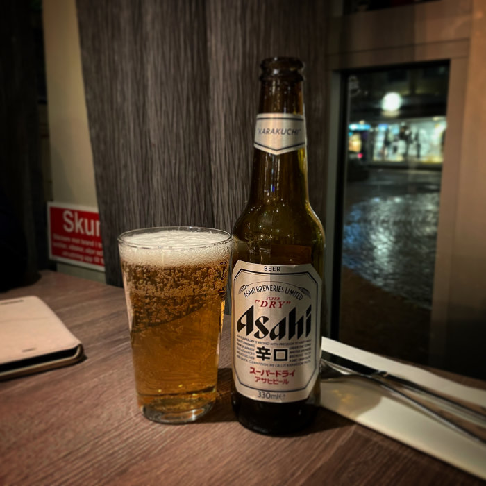Öppnad flaska Asahi-öl med fyllt glas framför, interiör med utsikt mot en nattlig gatuvy.