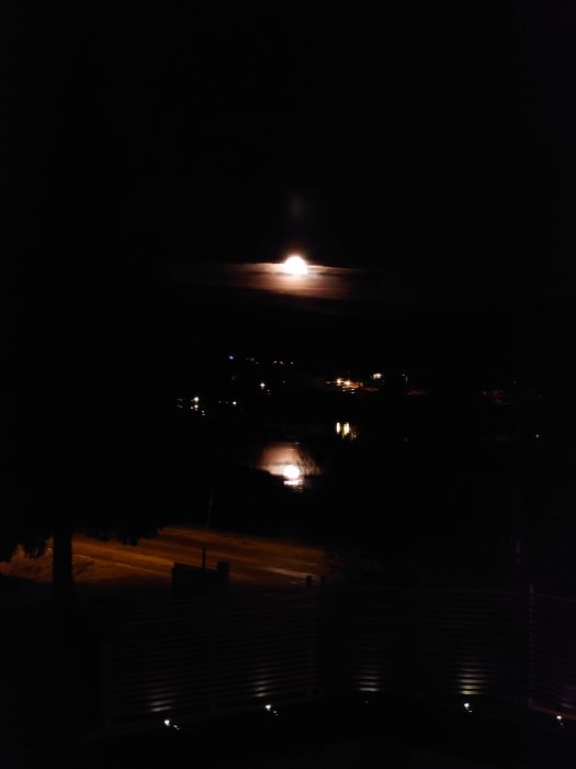 Nattbild med fullmåne bakom moln ovanför upplyst väg och vatten.