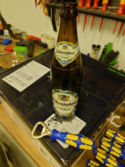 Öppnad ölflaska av märket Weihenstephaner Festbier på ett arbetsbord med verktyg och handskar.