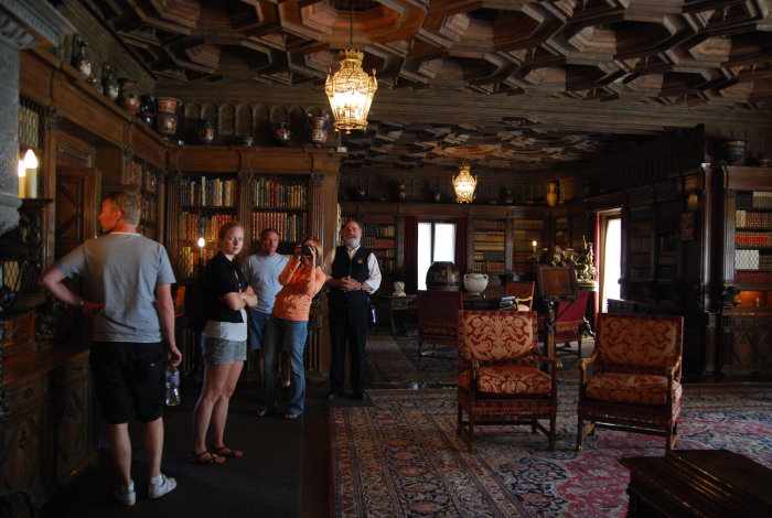Besökare i en storslagen sal med träpanel och tak, antika möbler och böcker, på Hearst Castle.