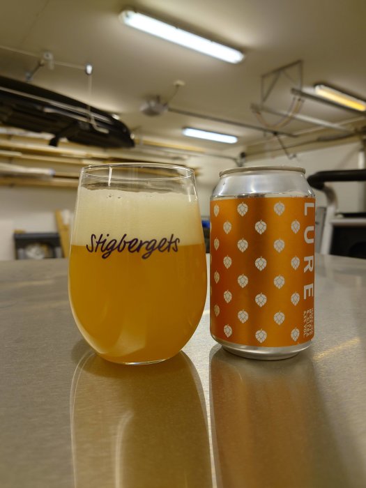 Ett glas öl med skum och en tillhörande burk på en bänk i ett verkstadsliknande utrymme.
