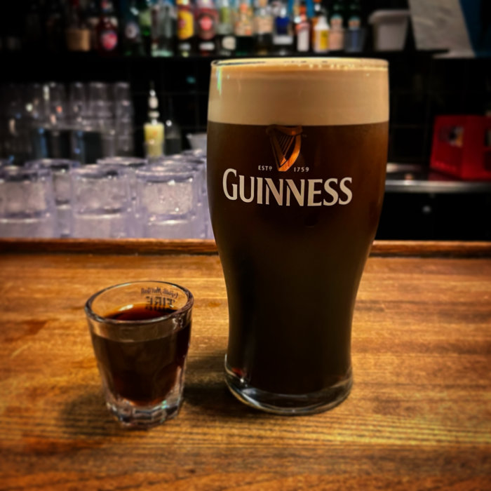 Ett pintglas Guinness öl med krämigt skum bredvid ett litet glas på en bardisk.