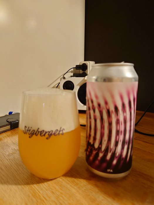 Ett glas öl med skum vid sidan av en ölburk med mönstrad etikett på ett bord, mikrofon i bakgrunden.