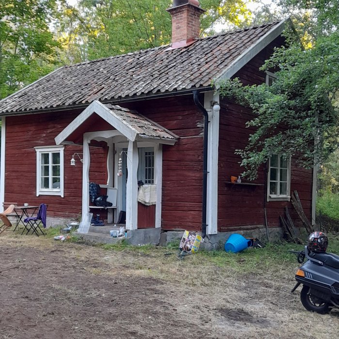 Rödmålat torp med slitna takpannor och diverse byggmaterial ute, samt en skoter parkerad framför.