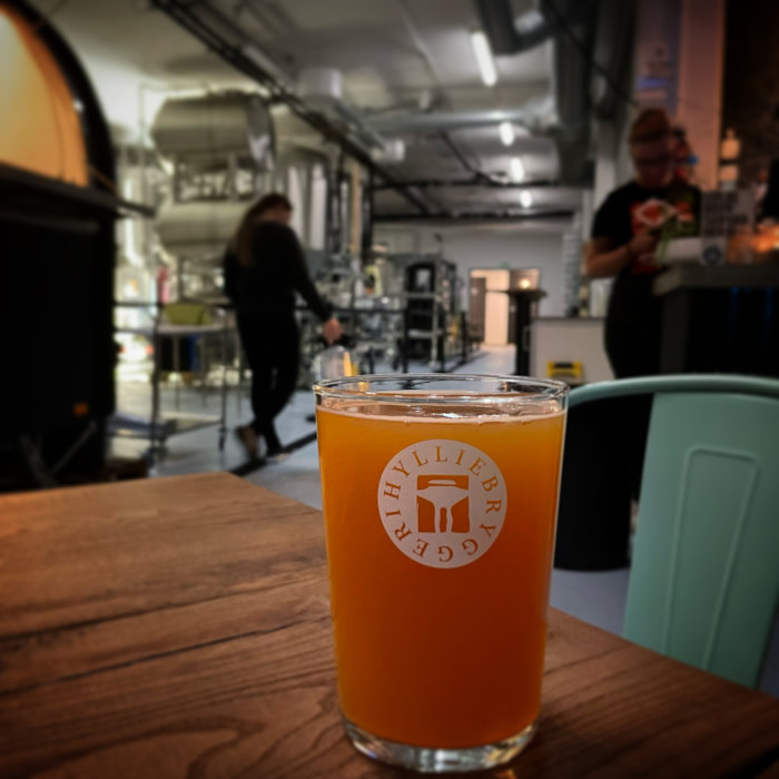 Ölglas med logotyp i förgrunden, suddig bakgrund av ett bryggeri med personer.