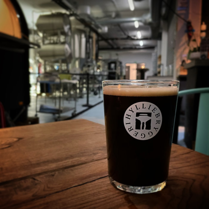 Ett glas mörk öl med skum på en bar med suddig bakgrund av bryggeriutrustning.