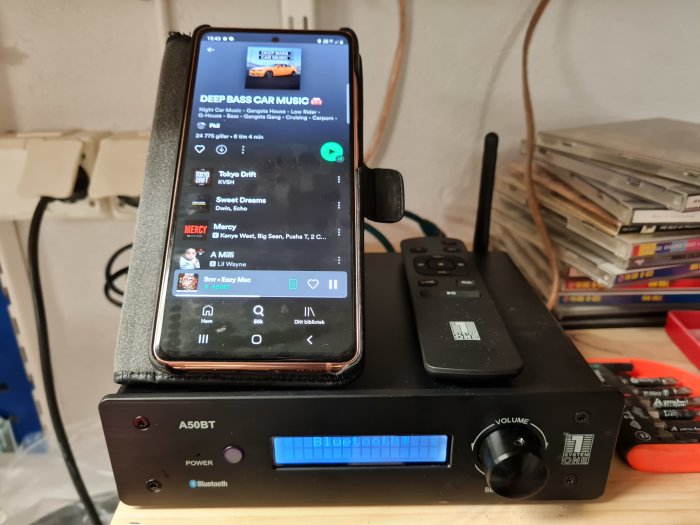 Mobiltelefon som spelar musik via en app, parkerad på en stereo med Bluetooth-funktion och en samling CD-skivor i bakgrunden.