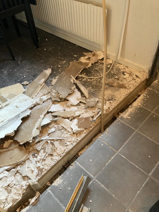 Rivning av gipsvägg med synlig regel och rörledning, spillror på golvet vid en radiator.
