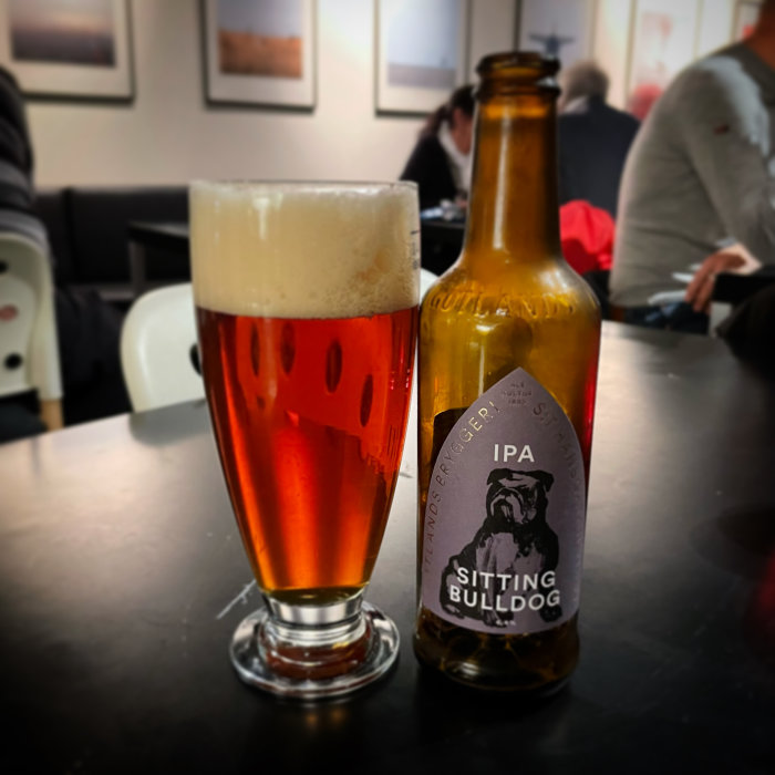 Ett glas med öl bredvid en tom IPA-flaska med etikett "Sitting Bulldog" på ett bord i ett café.