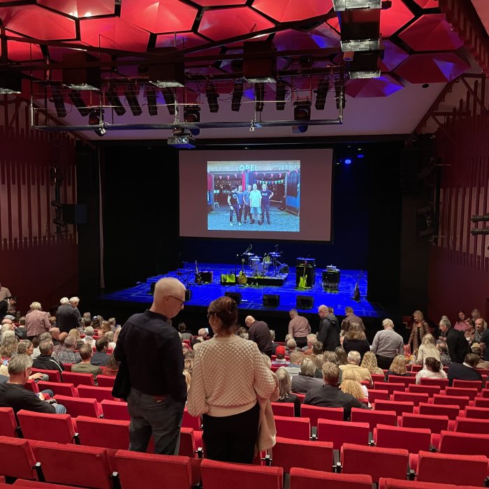 Publik samlad i en teatersalong med röda säten inför en föreställning med en tom scen och instrument.