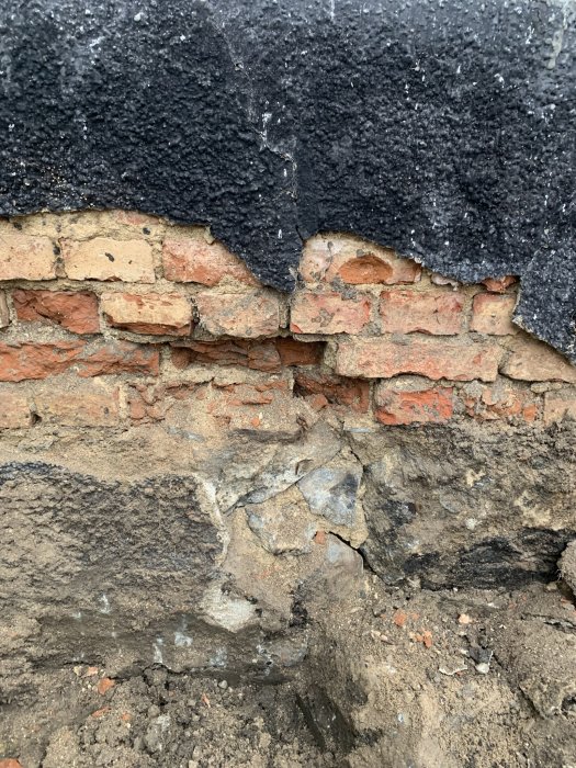 Skadad mur med flagnande svart tätskikt över röda tegelstenar och underliggande betongkonstruktion.