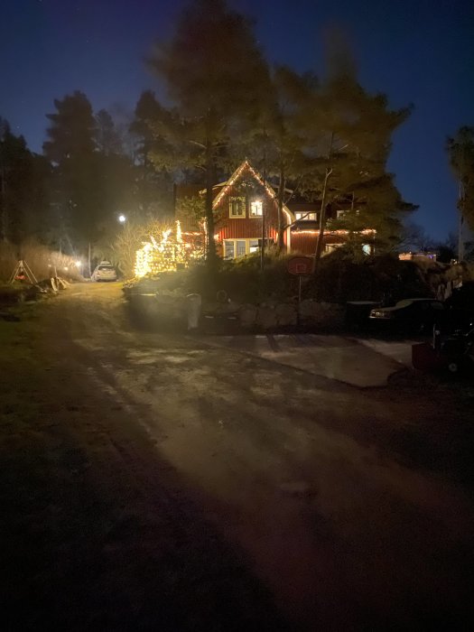 Ett hus med julbelysning under kvällstimmen, synligt upplyst träd och en del av uppfarten.