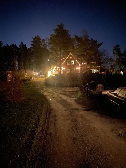 Kvällsbild av ett hus med julbelysning, omgivet av träd och en grusväg i förgrunden.