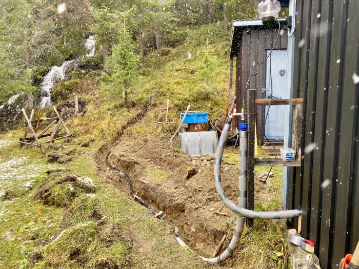 Vattenledning med PEM-slang och värmekabel som löper från damm till utsidan av en fjällstuga under snöfall.