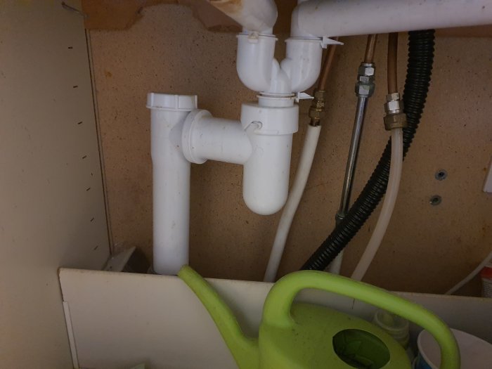 Undersidan av ett köksskåp med synliga rörkopplingar och vattenlås utan rengöringsöppning, intill en grön vattenkanna.