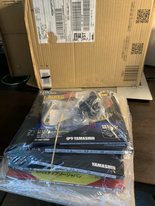 Ett öppnat paket från Amazon Japan med flera sågklingor och verktygskrok synligt genom plastförpackning.