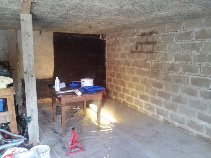 Oinrett garage med nakna betongväggar, träulltak och provisorisk arbetsbänk under renovering.