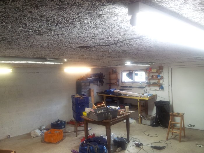 Ett rörigt garage med väggar och tak målade i vitt, lysrörsbelysning och verktyg på en arbetsbänk.