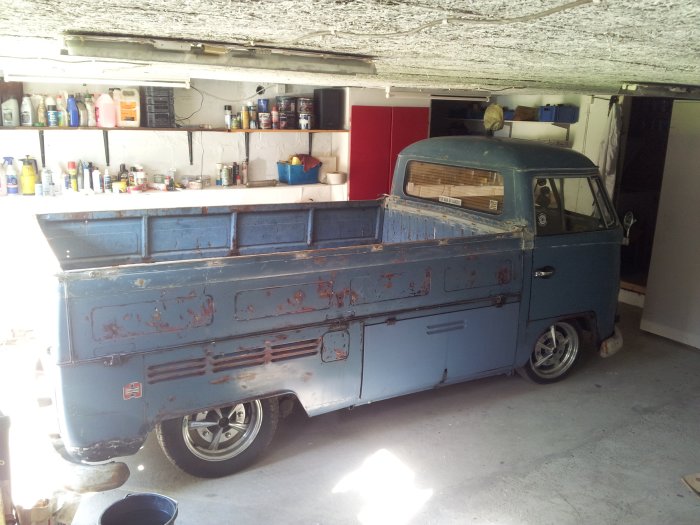 Blå pickup med patina parkerad i ett garage med målade vita väggar, träullstak och hyllor med verktyg och burkar.