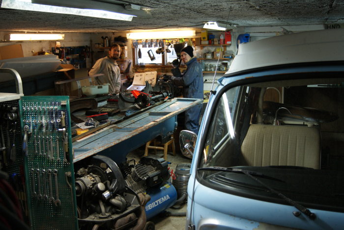 Två personer i ett trångt garage med verktyg, bil, arbetsbänk och oorganiserat kreativt kaos.